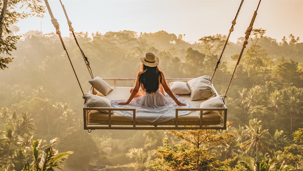 Photo of a young woman sitting on the swing.  Jungle Bed hanging over the tropical forest with Caucasian female resting while looking at the view, Bali, Indonesia. Rear view of a female sitting and enjoying the view.