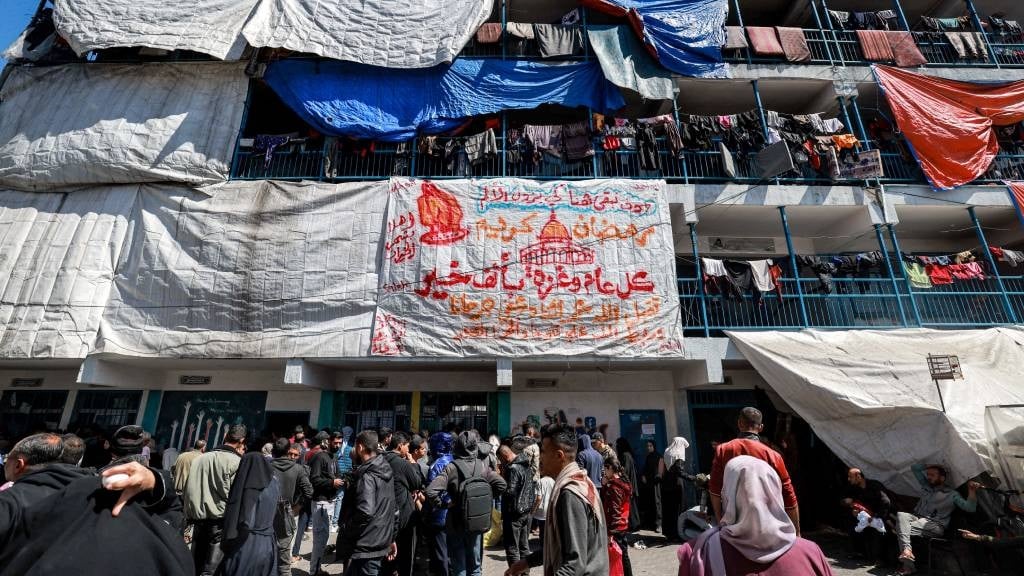 A large sheet displaying a greeting message for the Muslim holy month of Ramadan hangs from a building where below Palestinians gather to receive humanitarian aid, at a camp sheltering displaced Palestinians erected in a school run by the United Nations Relief and Works Agency for Palestine Refugees (UNRWA) in Rafah in the southern Gaza Strip on 13 March 2024, amid the ongoing conflict between Israel and the militant group Hamas. (Mohammed Abed/AFP)