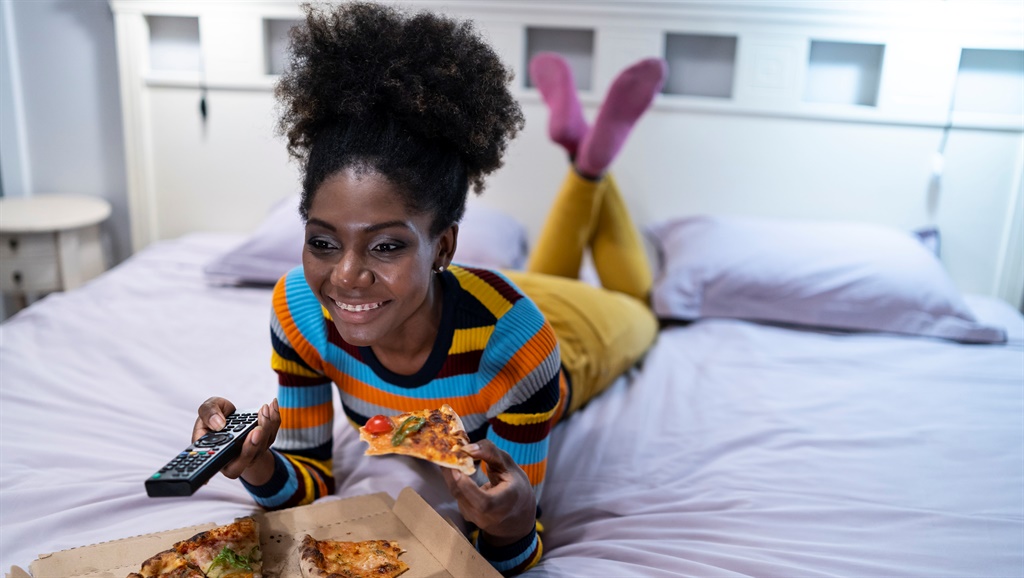 African woman lying down on bed, eating pizza and watching TV.