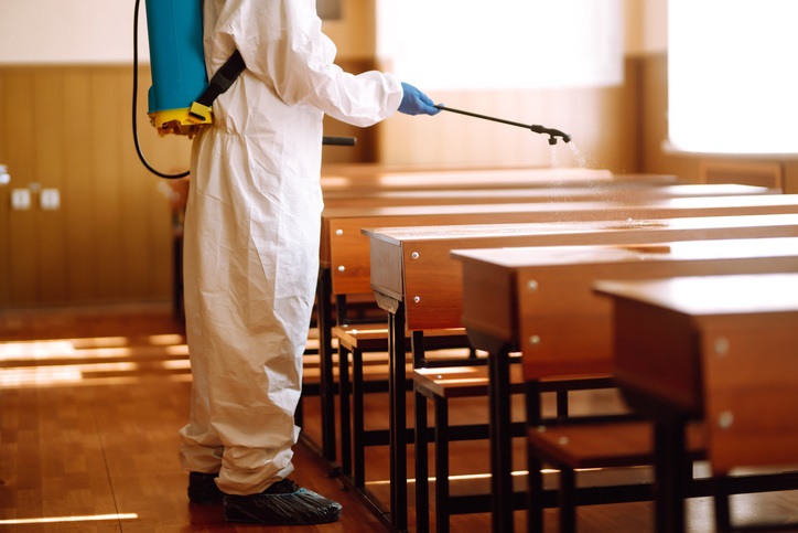 Man wearing protective suit disinfecting school class with spray chemicals to preventing the spread of coronavirus, pandemic in quarantine city. Disinfecting school desk. COVID-19.
