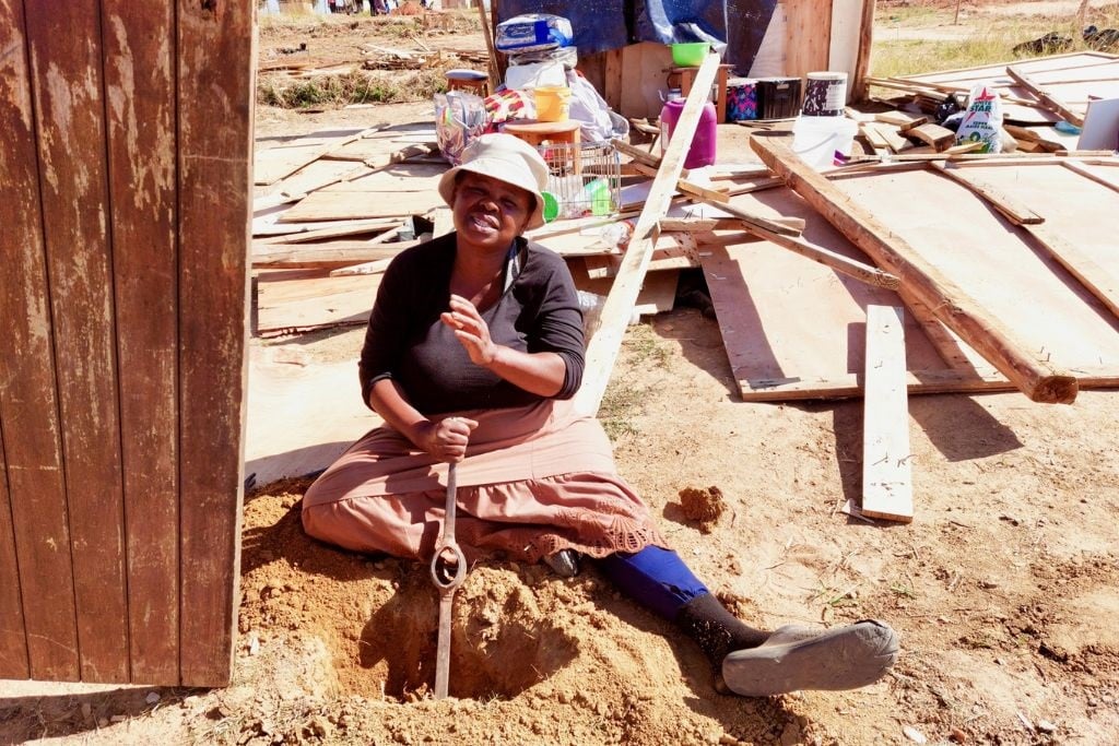 Tholakhele Nkosi digging a hole with just the head of a pickaxe to put up a support pole for her shack. (Kimberly Mutandiro/GroundUp)

