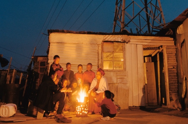 A family who live in a shack keep warm on a cold winter's evening around an outside fire. Their house, overshadowed by huge electric pylons, has no electricity.