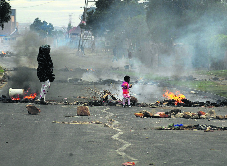 A mum and her toddler wander through burning tyres blocking the road leading to Ekuphumleni yesterday.      Photo by Chris Qwazi 