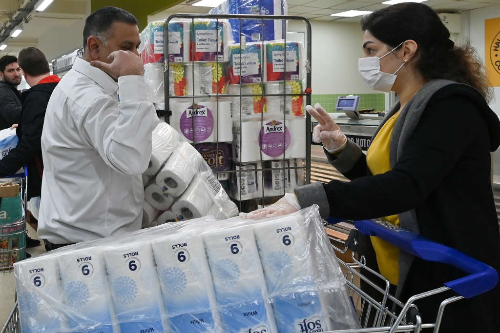 A woman wearing a mask gestures 'two' to confirm the limit set by this supermarket in London for toilet paper packs on 14 March 2020.