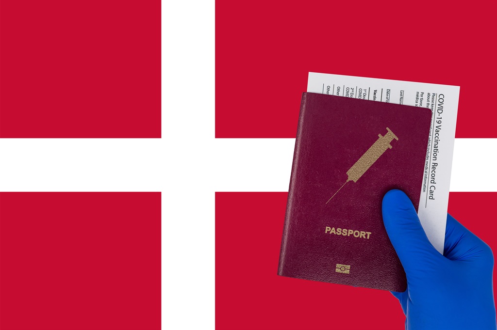 Hand with surgical glove holding a passport with syringe symbol and a vaccination record card or immunization certificate. Denmark flag on background