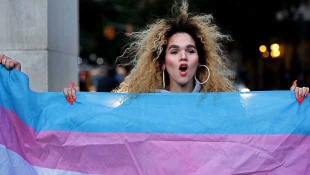 A woman of trans experience holds up the flag for Transgender and Gender Noncomforming people at a rally for LGBTQI+ rights at Washington Square Park