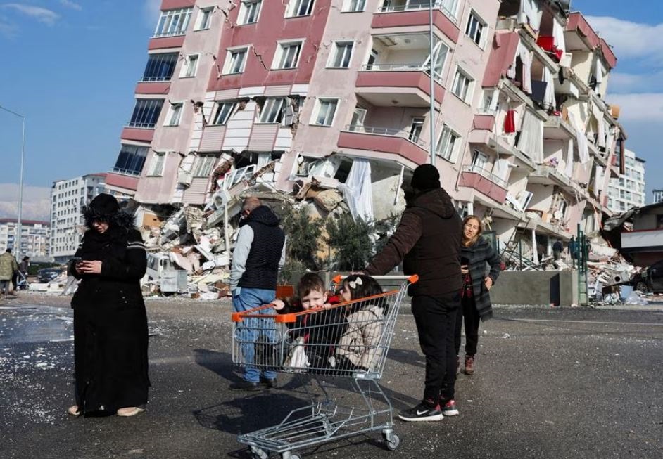 Children sit in a shopping cart near a collapsed building following an earthquake in Hatay, Turkey, February 7, 2023. 
