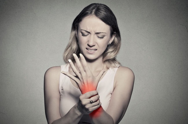 Young woman holding her painful wrist isolated on gray wall background. Sprain pain location indicated by red spot. Negative face expression .ÿÛ