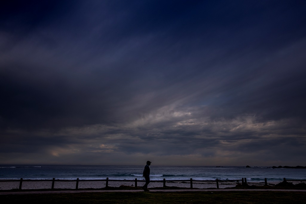 A man walks on footpath along Camps Bay beach as a cold front moves in over the ocean.