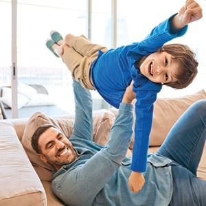 Father and son playing together on sofa in living room. Man lifting up his boy. Family staying at home due to pandemic COVID-19.